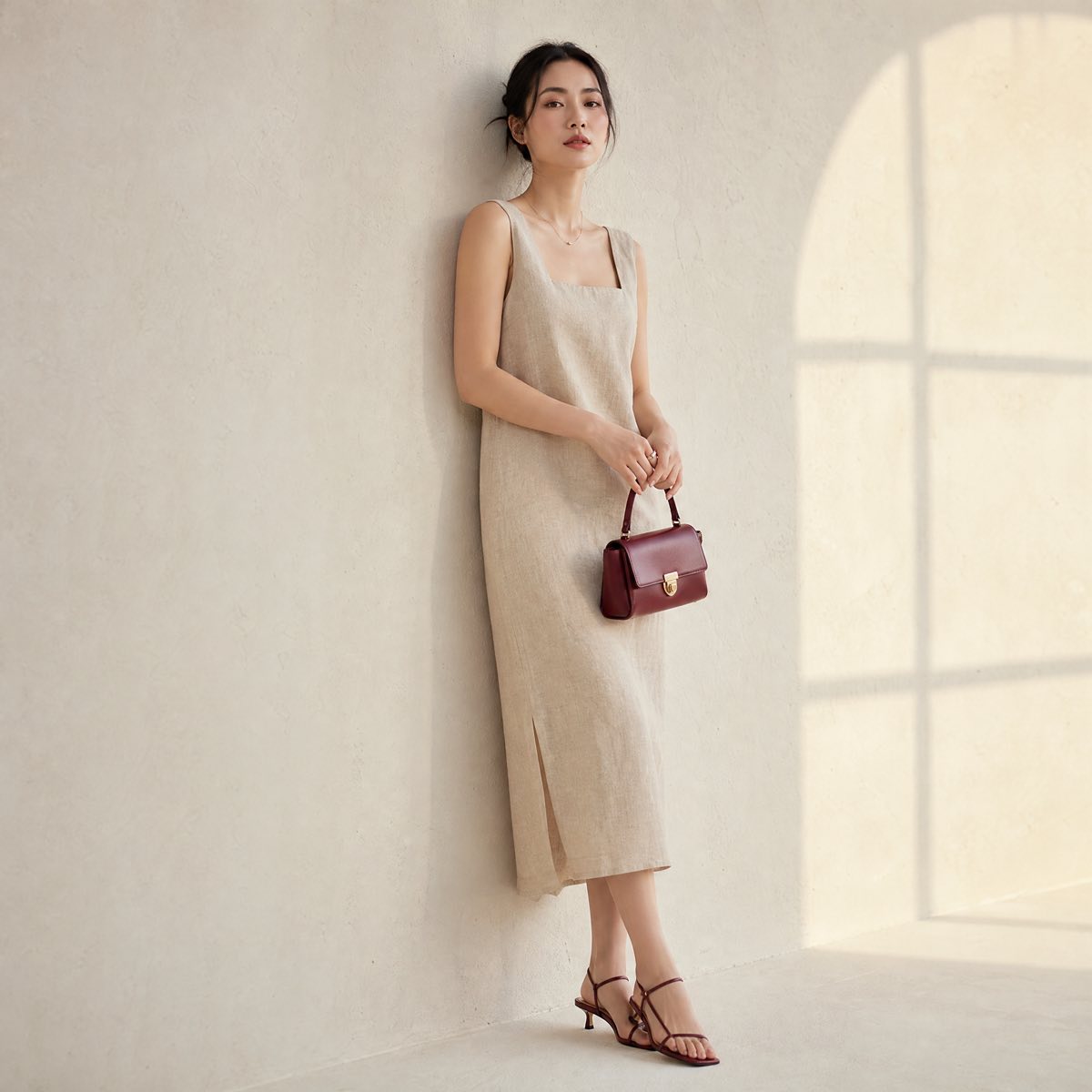 Người mẫu mặc đầm linen màu be của SƯƠNG Studio, đứng tựa tường trắng ấm với túi xách đỏ rượu vang